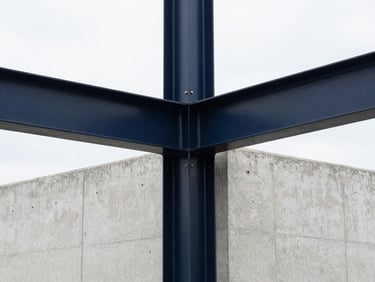 Close-up of structural steel joints in a blast-resistant concrete wall, sharp overcast daylight, high contrast, deep navy and stark white tones.