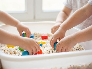 Toddlers playing with colorful sensory bins filled with rice and small toys, clean hands, bright natural window light, soft pastel tones, shallow depth of field, authentic moment