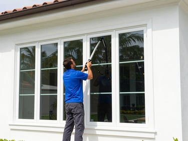 A technician in a vivid cobalt blue polo shirt and dark gray pants cleans large exterior windows of a luxury Florida home using a water-fed pole, bright Florida daylight.