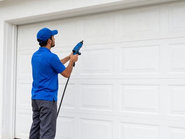 A technician in a vivid cobalt blue polo shirt and dark gray pants cleans a white garage door with a professional power washing wand, bright Florida daylight.