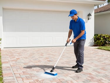 A technician in a vivid cobalt blue polo shirt and dark gray pants cleans a brick paver driveway with a professional surface cleaner, bright Florida daylight.