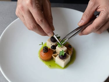 Overhead flat-lay of culinary preparation, hands of a chef using precision tweezers to place a single micro-herb on an architecturally structured dish, diffused natural light