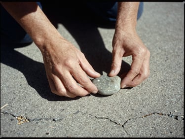 Cinematic 16mm film scan, high contrast, a close-up of hands placing a smooth river stone on a cracked concrete pavement, harsh sunlight, deep shadows