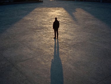 Anamorphic 35mm film still, high contrast, desaturated earth tones, a lone figure standing in a vast concrete plaza at dusk, golden light catching dust particles, quiet cinematic atmosphere