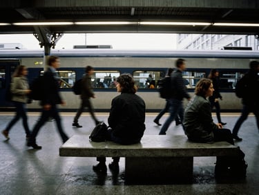 Anamorphic 35mm film still, a person sitting motionless on a concrete bench in a bustling train station, motion blur of crowds, high contrast