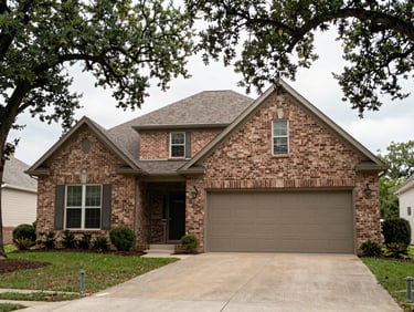 Tight exterior shot of a brick ranch-style home in a Houston suburb, wide driveway, two-car garage door, established oak trees framing the facade, bright overcast documentary light, no people