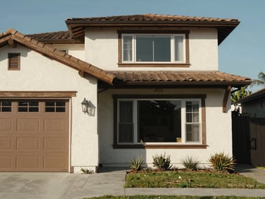 Close documentary shot of a craftsman-style home exterior in a Los Angeles residential neighborhood, stucco facade with wood trim, a garage door visible on the left, clear afternoon daylight, no people
