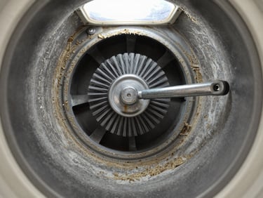 Close-up of a dryer vent interior showing compacted lint buildup along the duct walls, a metal cleaning brush mid-rotation inside the vent, bright daylight coming through the exterior vent cap, documentary framing