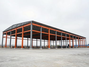 Wide-angle environmental shot of a massive newly constructed steel-frame warehouse, high contrast overcast sky, safety-orange structural beams visible, clean industrial concrete foreground.