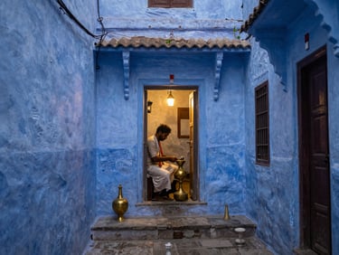 Narrow lane in Varanasi's old city at dawn, hand-painted blue walls, a brass craftsman working at an open doorway, warm lantern light spilling onto stone steps