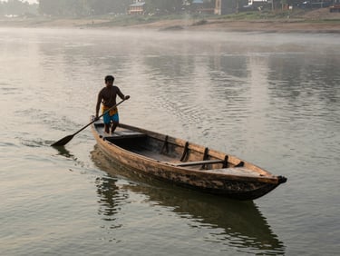 A wooden rowboat on the Ganga seen from slightly above, a local boatman at the oar, early morning mist on the water, the far bank faintly visible, documentary natural light