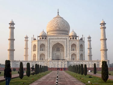 Wide shot of the Taj Mahal at dawn from the eastern garden path, soft diffused light, a lone visitor in the left foreground providing scale, mist on the Yamuna visible beyond the structure