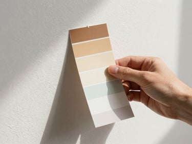 Close-up of a painter's hand holding a paint chip swatch against a wall surface in natural daylight, the wall texture visible, color sample centered in frame with crisp edges