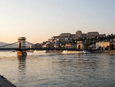 Morning light over the Danube at Budapest — the Chain Bridge in the mid-distance, a river ship passing beneath it, soft golden reflections on calm water, wide environmental framing with the Buda castle hill on the right