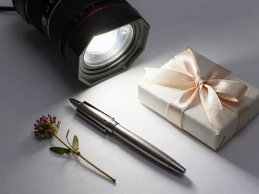 Curated gift set flat-lay: engraved pen, small floral stem, and ribbon-tied box on white surface, overhead studio light