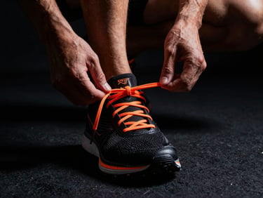 Dynamic close-up of an athlete preparing running shoes, sharp safety-orange laces, dramatic high-contrast studio lighting, deep black background