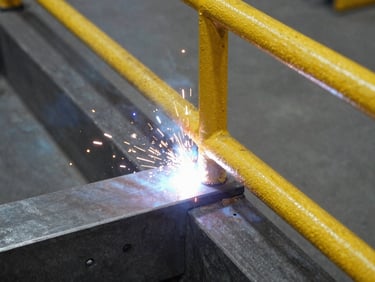 Close-up of industrial welding on a yellow steel safety handrail, bright sparks flying, sharp focus on the weld joint, industrial workshop setting