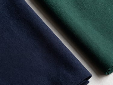 Overhead flat-lay of folded bolts of modest-fashion textile fabric — deep navy and forest green tones — on a plain light surface under natural daylight, fabric texture clearly visible, no garments, no styling