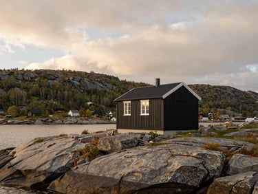 Architectural wide angle of a minimalist black timber cabin on a rocky Norwegian fjord edge, dramatic cloudy sky, soft daylight