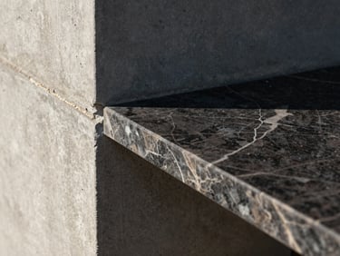 Close-up detail crop of a dark marble slab meeting a raw concrete wall, sharp sunlight highlighting the precise joint line, landscape orientation.