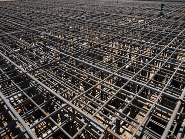 Low-angle perspective of a massive foundation steel cage layout, harsh midday sun casting sharp geometric shadows through the steel mesh.