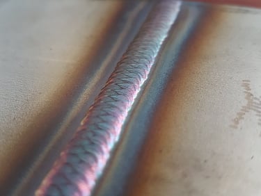 A precise TIG weld bead on stainless steel showing colorful heat tint and uniform ripples.