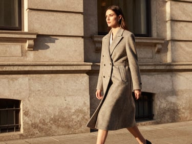 A sophisticated woman in a tailored wool coat walking past classic stone urban architecture, warm golden-hour light