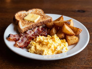 A hearty breakfast plate with fluffy scrambled eggs, thick-cut smoked bacon, golden hash browns, and buttered sourdough toast under soft morning light.
