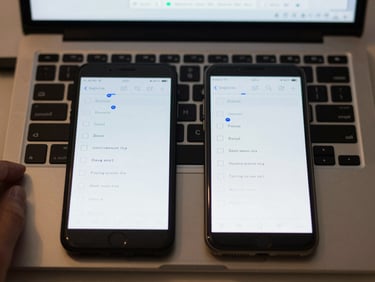 Close-up overhead view of a laptop keyboard and a smartphone placed side by side on a light desk surface, both screens displaying a simple multiple-choice interface, ordinary overhead classroom light, no dramatic shadows, hands partially visible at keyboard edges