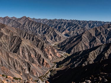 Wide panoramic Andes mountain vista from a high viewpoint, layered ridgelines receding into the distance under a deep blue midday sky, valley floor visible far below, minimal vegetation, geological scale dominant