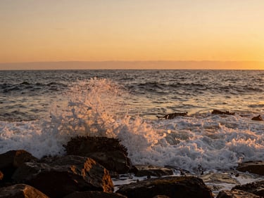 Wide coastal shot of Viña del Mar at golden hour, waves breaking against dark rock in the foreground, warm orange light on the water, low horizon, no people