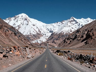 Wide environmental shot looking east along a straight Andean valley road at midday, massive snow-covered mountain ridgeline filling the horizon, ochre rock walls on either side, no vehicles, deep blue sky