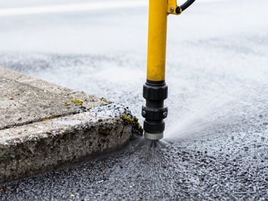 Close-up of industrial pressure washing equipment stripping away heavy grime and moss from a concrete curb, bright overcast daylight, high contrast