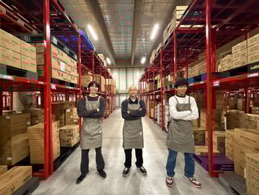 株式会社ロジ・クム提供の物流倉庫内の保管棚の通路でエプロン姿のスタッフ3人が立つ様子|堺市のニュースならサカイタイムズ