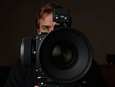 Moody portrait of a film director behind an anamorphic camera lens, low-key dramatic chiaroscuro lighting, rich contrast, 35mm