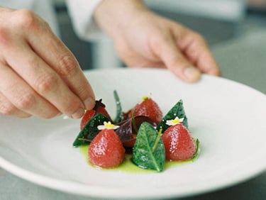 Close-up macro shot of a chef's hands plating a vibrant dish in a softly lit kitchen, focus on the texture of the food, warm editorial photography, soft natural daylight, rich crimson and forest green tones, shallow depth of field, 35mm film.
