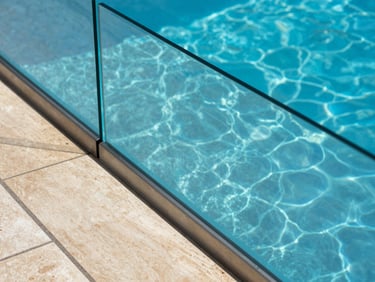 Overhead close-up of a pool perimeter glass railing in bright Andalusian sunlight, blue water visible through the transparent panel, stainless steel base channel flush with the pool coping, no people