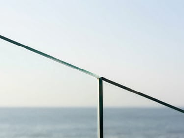 Wide macro shot of laminated glass cross-section held up to coastal daylight, the interlayer film visible as a thin translucent band between two clear panes, bright white sky behind, clean minimal composition, no people