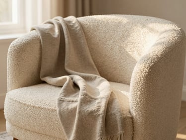 Close-up of a textured cream bouclé armchair paired with a draped neutral linen throw blanket, soft warm lighting