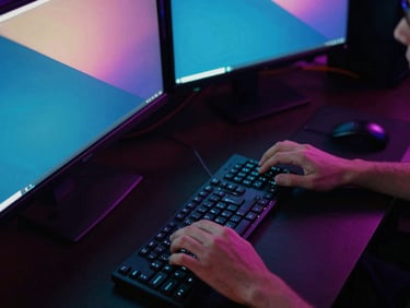 High-angle view of a game developer working at a dual-monitor setup in a low-key ambient room, neon screen glow casting onto hands and keyboard.