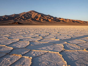 Wide environmental shot of the Atacama salt flats at golden hour, cracked earth surface stretching toward distant rust-colored mountains, long shadows crossing the foreground, no people