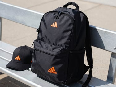 Close-up of an Adidas black backpack and cap on an outdoor metal bench, bright natural sunlight