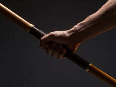 Dramatic close-up of hands gripping a dark wooden Bo staff, side-lit with warm gold highlights, dark charcoal background