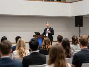 Corporate conference room mid-event, keynote speaker at podium bathed in stage light, audience in soft focus, clean architectural venue, event documentation framing
