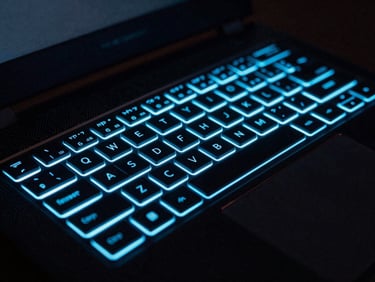 Close-up of a high-performance gaming laptop with RGB keyboard glowing electric blue, on a dark surface with subtle circuit board patterns, neon-lit hardware photography, deep obsidian background, electric cyan and blue rim lighting, macro lens, high contrast, 35mm
