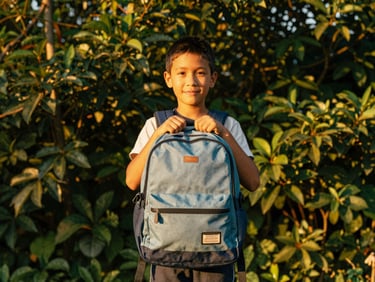 Child proudly showing a new backpack and school supplies, warm documentary photography, natural golden-hour sunlight, eye-level shot, authentic emotional expressions, rich forest green and gold tones, 35mm lens
