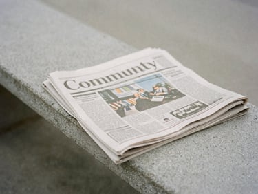 Close-up of a printed community newspaper on a concrete bench, soft overcast daylight, muted desaturated tones, quiet editorial mood.