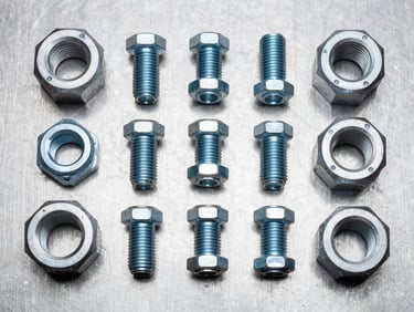 Overhead flat-lay of industrial bolts, flanges, and pipe fittings arranged on a steel surface under studio strobe lighting, cool white light, sharp detail on threading and surface finish