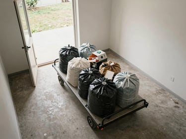 Overhead environmental shot of a cleared room with a flat-bed dolly loaded with bagged debris near an open exterior door, natural daylight flooding in from outside, honest before-clearance framing