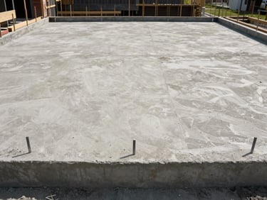 Straight-on daylight shot of a finished concrete residential foundation and ground-floor slab — formwork removed, level surface visible, construction stakes still in place at perimeter